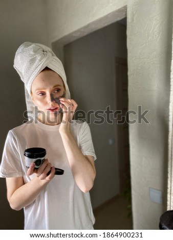 beautiful woman in a towel on her head puts a clay gray mask on her face while looking in the mirror. Beauty treatments at home.