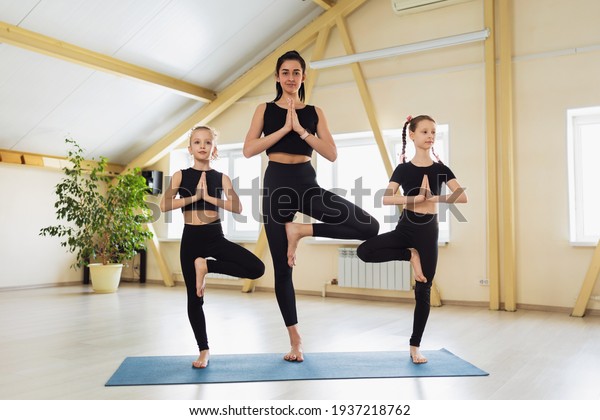 Beautiful\
woman trainer with two students in black sportswear, practicing\
yoga, perform Vrikshasana exercise on a gymnastic mat, tree pose\
with namaste, exercising in a yoga\
studio.