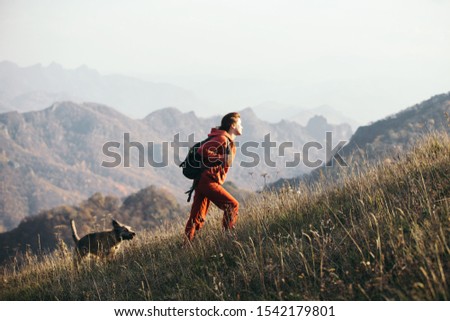 Beautiful woman traveler climbs uphill with a dog on a background of mountain views. She is with a backpack and in red clothes.