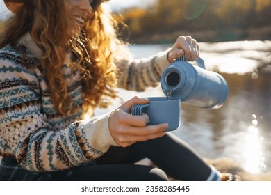 Beautiful woman traveler in a hat sits on a log near the river, drinks a hot drink from a thermos. Smiling female tourist enjoys the autumn landscape near the river. The concept of travel, relax.