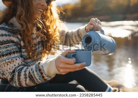 Beautiful woman traveler in a hat sits on a log near the river, drinks a hot drink from a thermos. Smiling female tourist enjoys the autumn landscape near the river. The concept of travel, relax.
