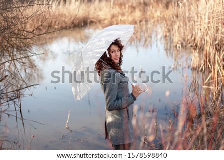 Beautiful woman with umbrella walking outdoor at the nature. Female wearing grey stylish coat and posing at background lake and grass on autumn or early spring day