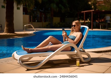 Beautiful woman uses her phone while sitting on a lounger by the pool during her holiday. European woman sitting at the swiming pool and using the smart phone to check here work email or social media. - Powered by Shutterstock