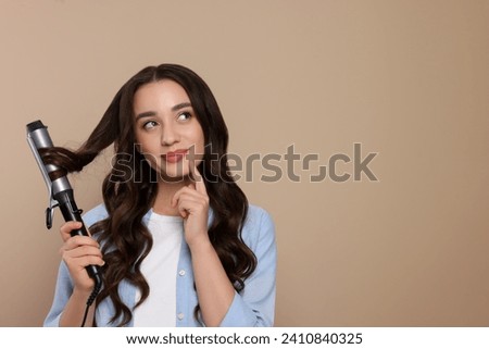 Beautiful woman using curling hair iron on beige background. Space for text