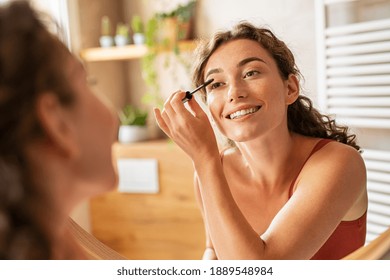 Hermosa mujer con rímel en pestañas en el baño por la mañana. Joven sonriente aplicando maquillaje ocular y mirando al espejo. Chica de belleza aplicando rímel negro en el baño de su casa.