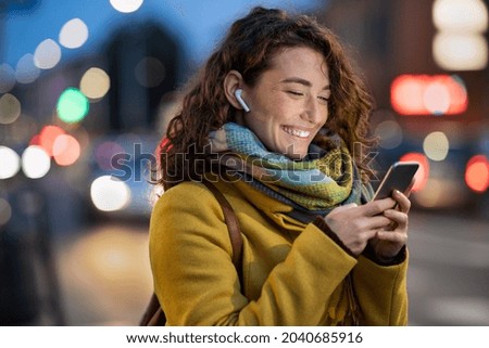 Beautiful woman using mobile phone while listening voice note with wireless earbuds. Smiling girl changing songs from smart phone in a city street. Happy smiling woman chatting on smartphone in winter