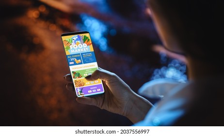 Beautiful Woman Using Smartphone For Ordering Food Via Online Food Market Store While Walking Through Night City Street Full Of Neon Light. Over The Shoulder Close-up Shot Of A Phone Screen