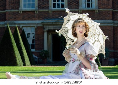 Beautiful woman in vintage clothing sitting on lawn in front of stately home holding parasol and drinking sparkling wine