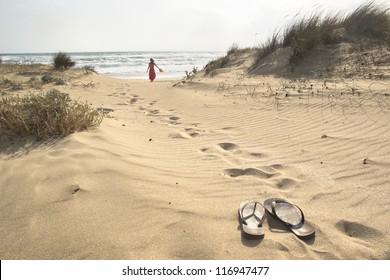 A beautiful woman walks barefoot through sand dunes towards to sea leaving her flip flops behind