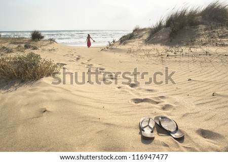 A beautiful woman walks barefoot through sand dunes towards to sea leaving her flip flops behind