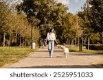 A beautiful woman in warm autumn clothing strolls through a peaceful park with her dog.