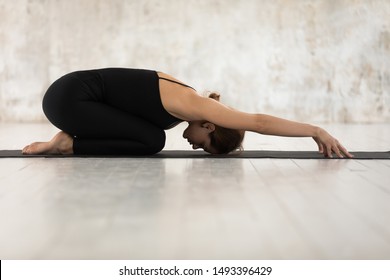 Beautiful woman wearing black sportswear practicing yoga, relaxing in Child pose, Balasana exercise, sporty girl working out, stretching at home or in yoga studio with grey walls