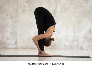 Beautiful woman wearing black sportswear practicing yoga, doing uttanasana exercise, standing forward bend pose, attractive sporty girl working out at home or in yoga studio with grey walls