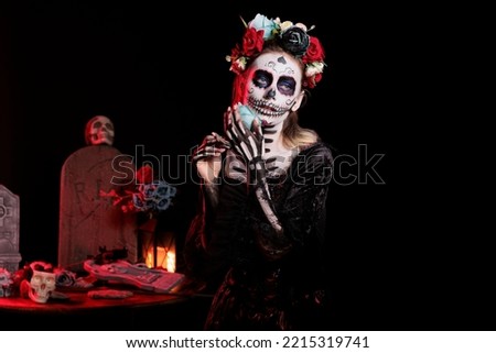 Beautiful woman wearing black and white make up and flowers crown to cleebrate mexican halloween ritual. Goddess of death with horror carnival costume and body art, holding rose.