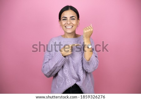 Beautiful woman wearing a casual violet sweater over pink background smiling and pointing his watch because it's late