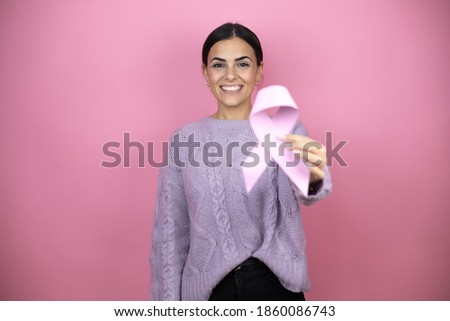 Beautiful woman wearing a casual violet sweater over pink background holding a cancer ribbon with a confident smile showing teeth
