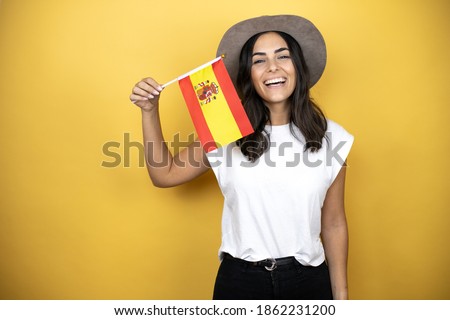 Beautiful woman wearing casual white t-shirt and a hat standing over yellow background holding the spanish flag