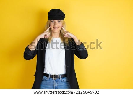 Beautiful woman wearing casual white t-shirt and a cap over isolated yellow background smiling confident showing and pointing with fingers teeth and mouth