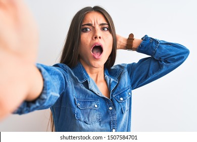 Beautiful woman wearing denim shirt make selfie by camera over isolated white background Crazy and scared with hands on head, afraid and surprised of shock with open mouth