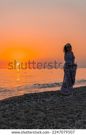 Beautiful woman wearing dress and sunglasses standing at beach with scenic view of seascape and clear sky in the background during sunset