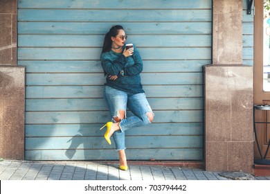 Beautiful woman wearing fall sweater, ripped jeans and colorful shoes drinking take away coffee standing against cafe wall on city street. Casual fashion, elegant everyday look. Plus size model.