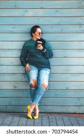 Beautiful woman wearing fall sweater, ripped jeans and colorful shoes drinking take away coffee standing against cafe wall on city street. Casual fashion, elegant everyday look. Plus size model.