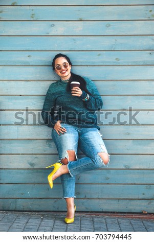Beautiful woman wearing fall sweater, ripped jeans and colorful shoes drinking take away coffee standing against cafe wall on city street. Casual fashion, elegant everyday look. Plus size model.