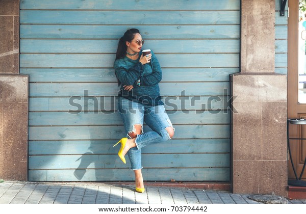 秋のセーターを着た美しい女性 リップしたジーンズ カラフルな靴を履いた女性が 街の通りのカフェの壁に立ってコーヒーを飲む カジュアルファッション エレガントな日常の外観 プラスサイズモデル の写真素材 今すぐ編集