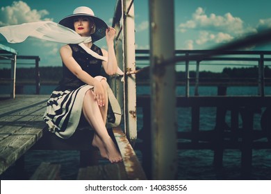 Beautiful woman wearing hat and white scarf sitting on old wooden pier