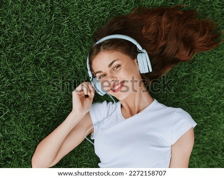 Beautiful woman wearing headphones listening to music lying on the grass on the lawn in the park in spring and smiling with teeth