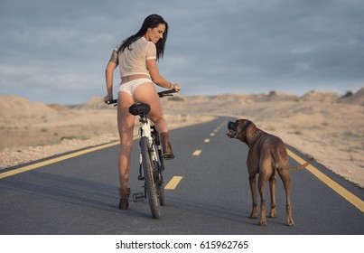 A Beautiful Woman Wearing A Short Blouse And Laced Panties And Boots With Tattoos Riding A Mountain Bike On A Deserted Road With Her Boxer Dog With A Desert Background 