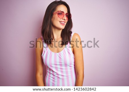 Beautiful woman wearing striped pink swimsuit and heart shaped sunglasses over pink background looking away to side with smile on face, natural expression. Laughing confident.