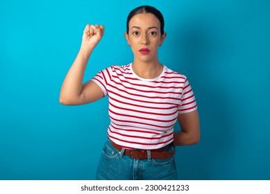 beautiful woman wearing striped T-shirt over blue studio background feeling serious, strong and rebellious, raising fist up, protesting or fighting for revolution.