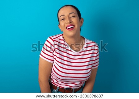 beautiful woman wearing striped T-shirt over blue studio background with broad smile, shows white teeth, feeling confident rejoices having day off.