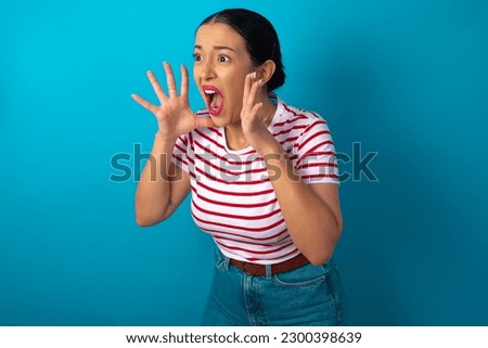 beautiful woman wearing striped T-shirt over blue studio background with shocked facial expression, holding hands near face, screaming and looking sideways at something amazing.
