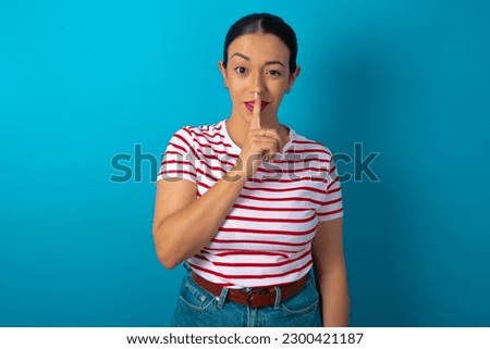 beautiful woman wearing striped T-shirt over blue studio background makes hush gesture, asks be quiet. Don't tell my secret or not speak too loud, please!