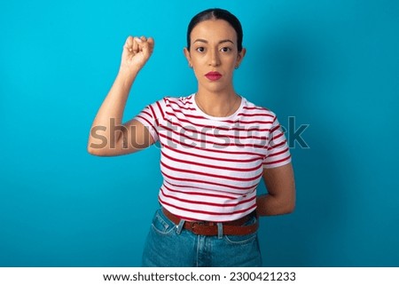 beautiful woman wearing striped T-shirt over blue studio background feeling serious, strong and rebellious, raising fist up, protesting or fighting for revolution.