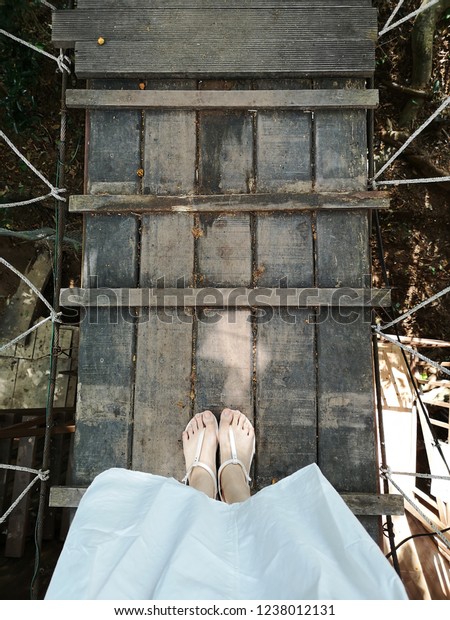 Beautiful Woman Wearing White Skirt or Dress\
and Nude Sandals on Brown Wood. Female is Legs and Shoes on Wooden\
Bridge covered by Rope in Autumn\
Background