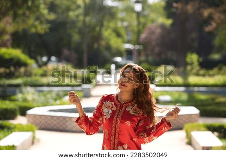 A beautiful woman wears a traditional Moroccan dress in red and embroidered in gold and silver. The girl raises her arms for wedding photos in a Moroccan style garden with fountains and lush greenery.