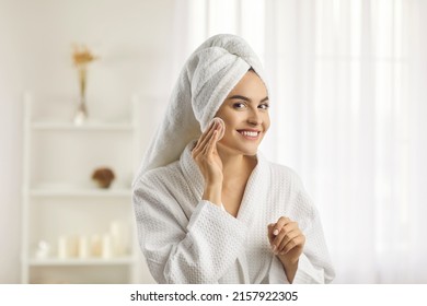 Beautiful woman in white bathrobe and towel turban on head looking at reflection in mirror and putting toner on face with cotton disk pad preparing herself for new day. Beauty and skin care concept - Powered by Shutterstock