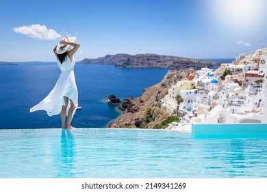 A beautiful woman in a white dress stands by the swimming pool and enjoys the view over the village of Oia, Santorini island, Greece, during her summer vacation time