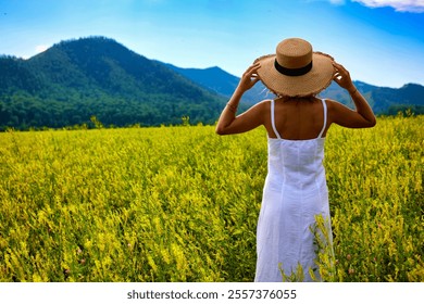 A beautiful woman in a white dress and straw hat stands in a golden mustard field, with a clear blue sky above, creating a serene and picturesque outdoor scene. - Powered by Shutterstock