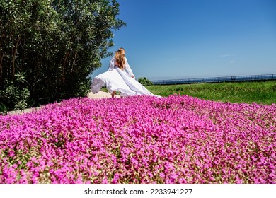 a beautiful woman in a white flowing long dress stay near a beautiful field with pink flowers rear view - Shutterstock ID 2233941227