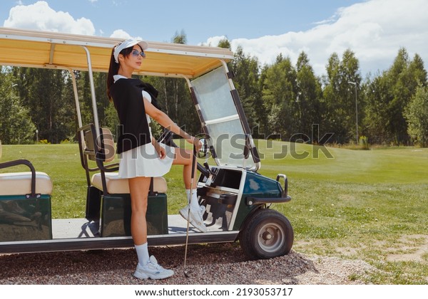 Beautiful woman in white outfit standing near\
golf cart. Rich\
lifestyle