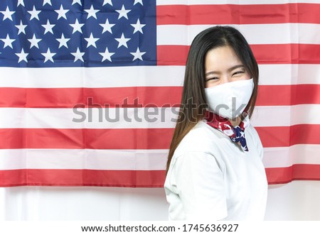 A beautiful woman in white shirt wearing mask to protect virus and posing with background of american flag for celebrating national independence day on 4th of July