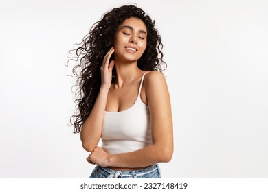 beautiful woman in a white T-shirt and blue jeans with long curly hair stands against a white background and smiles with her eyes closed in pleasure - Powered by Shutterstock