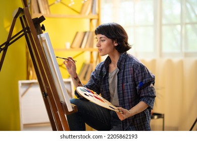 A Beautiful Woman Who Is An Artist And Has Paint Over Her Hands Is Working On Painting Something On A Large Canvas In Front Of Her While She Is Sitting On A Stool