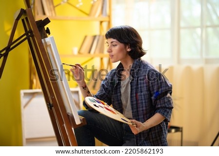 A beautiful woman who is an artist and has paint over her hands is working on painting something on a large canvas in front of her while she is sitting on a stool