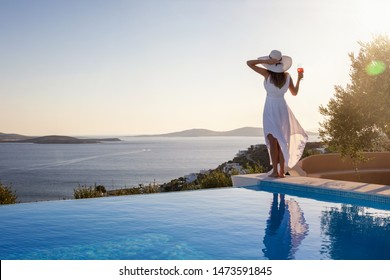 Beautiful Woman In Whte Dress And Hat Stands By The Pool Edge And Enjoys The Summer Sunset With A Glass Of Wine And View To The Mediterranean Sea