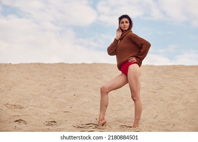 Beautiful Woman With Windy Hair Standing On Sandy Beach. Stylish Young Sexy Female In Knitted Sweater And Bare Legs Relaxing On Coast.